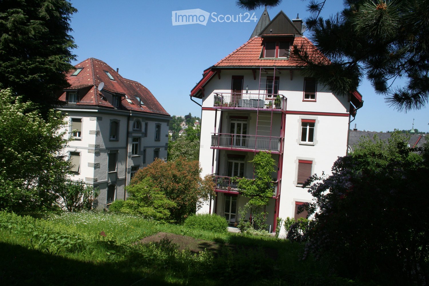 Two-story residential buildings with red-tiled roofs, white walls, and multiple windows. One building features a balcony with railings and potted plants.