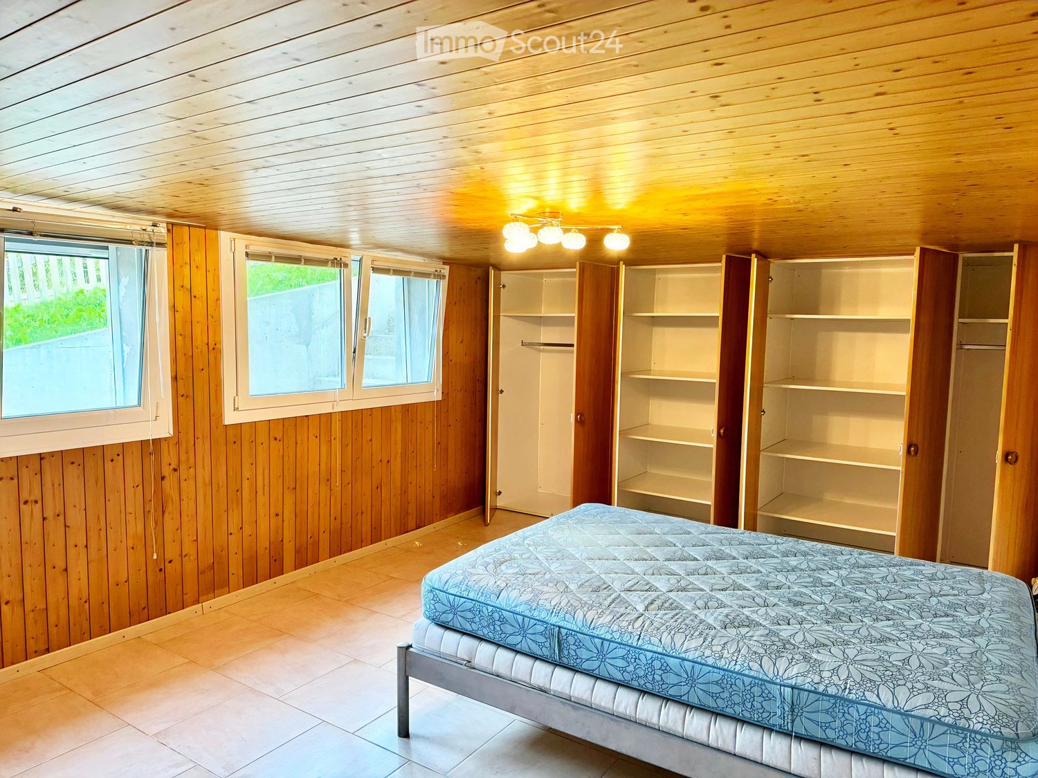 Bright bedroom with wooden walls and ceiling, large windows, a single bed with blue patterned bedding, and an open wardrobe with empty shelves.