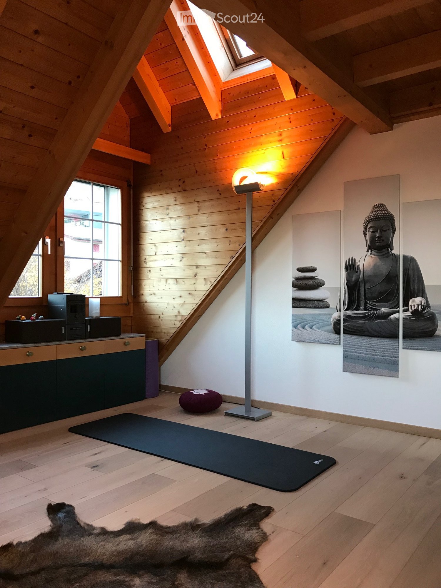 A spacious, well-lit attic room with wooden walls and ceiling beams, featuring a yoga mat, a floor lamp, a cabinet, a Buddha statue, and a stack of stones.