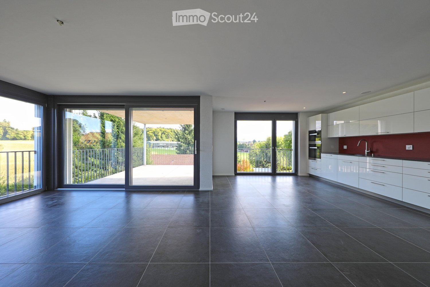 Modern, open-plan kitchen and living area with dark tiled flooring, white cabinets, a red backsplash, and large sliding glass doors leading to a balcony.