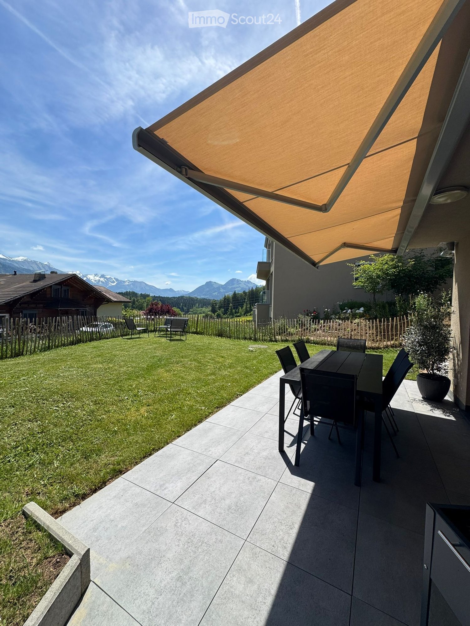 Outdoor patio with a large retractable awning, a dining table with chairs, and a view of a green lawn and mountains in the background.