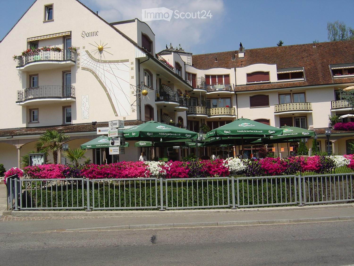 Multi-story building with balconies, green umbrellas, and colorful flowers in front.