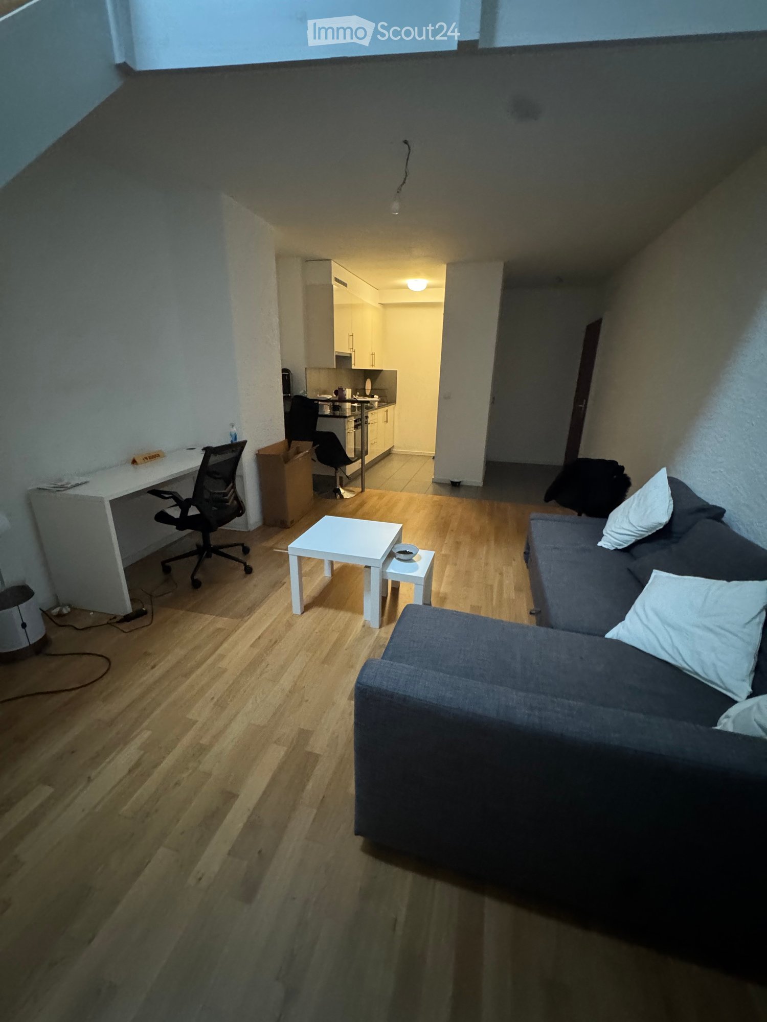 A modern, open-plan living and working space with wooden flooring, a grey sofa, a small white coffee table, a desk with a chair, and a small kitchen area in the background.