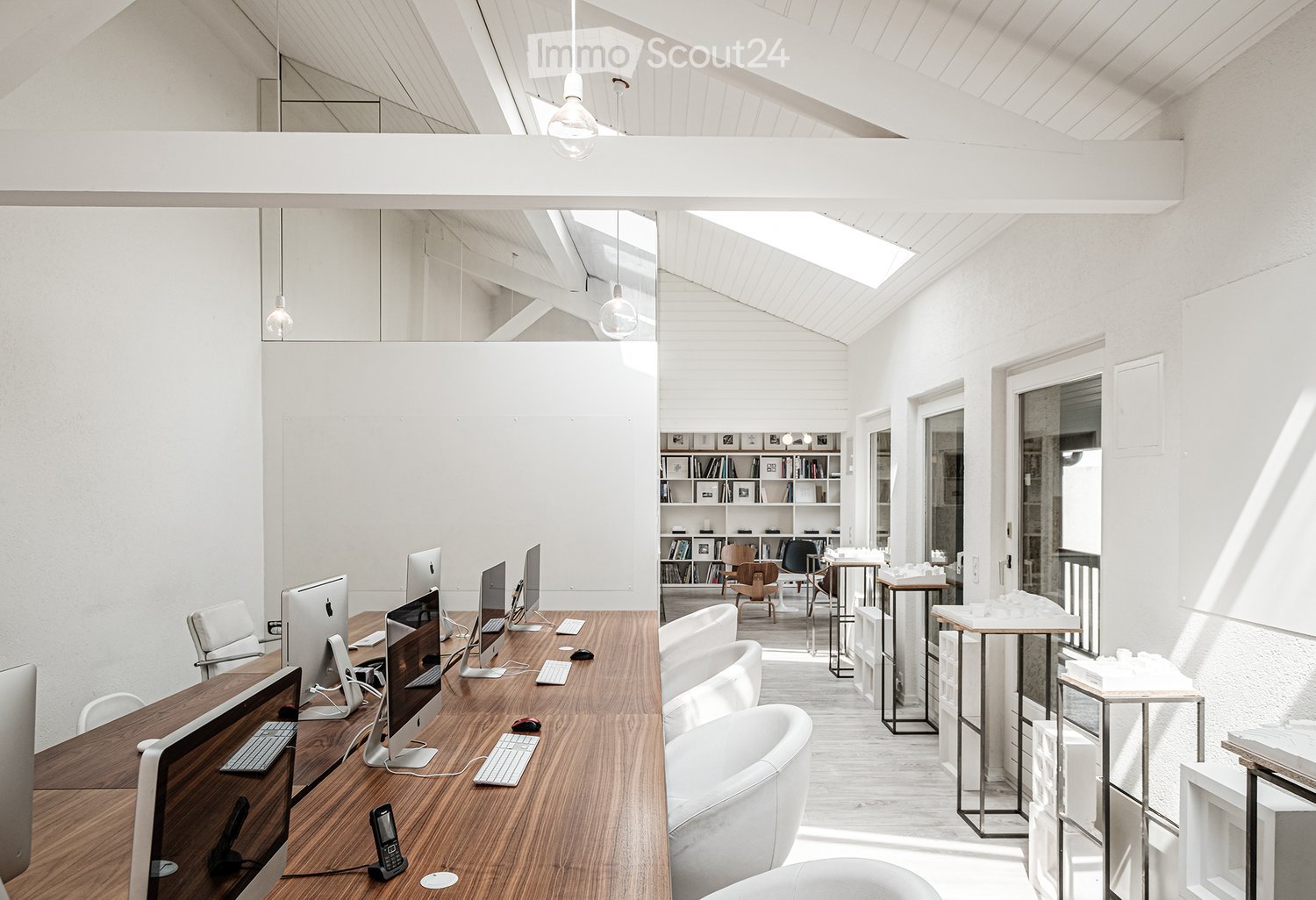 Bright, open-plan office space with wooden desk, multiple computer screens, ergonomic chairs, and a bookshelf in the background.