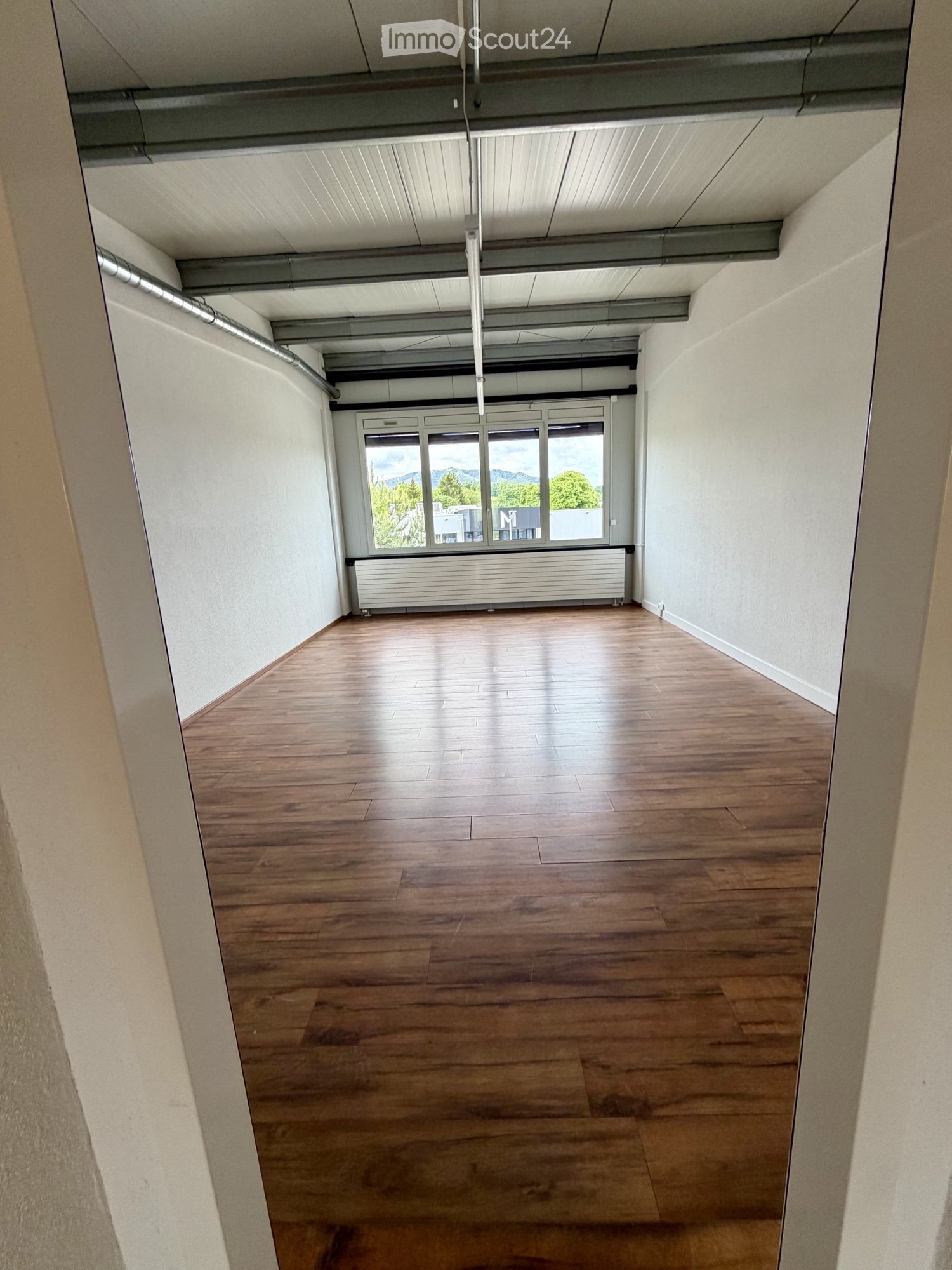 Empty room with wooden flooring, white walls, large windows, and exposed ceiling beams.