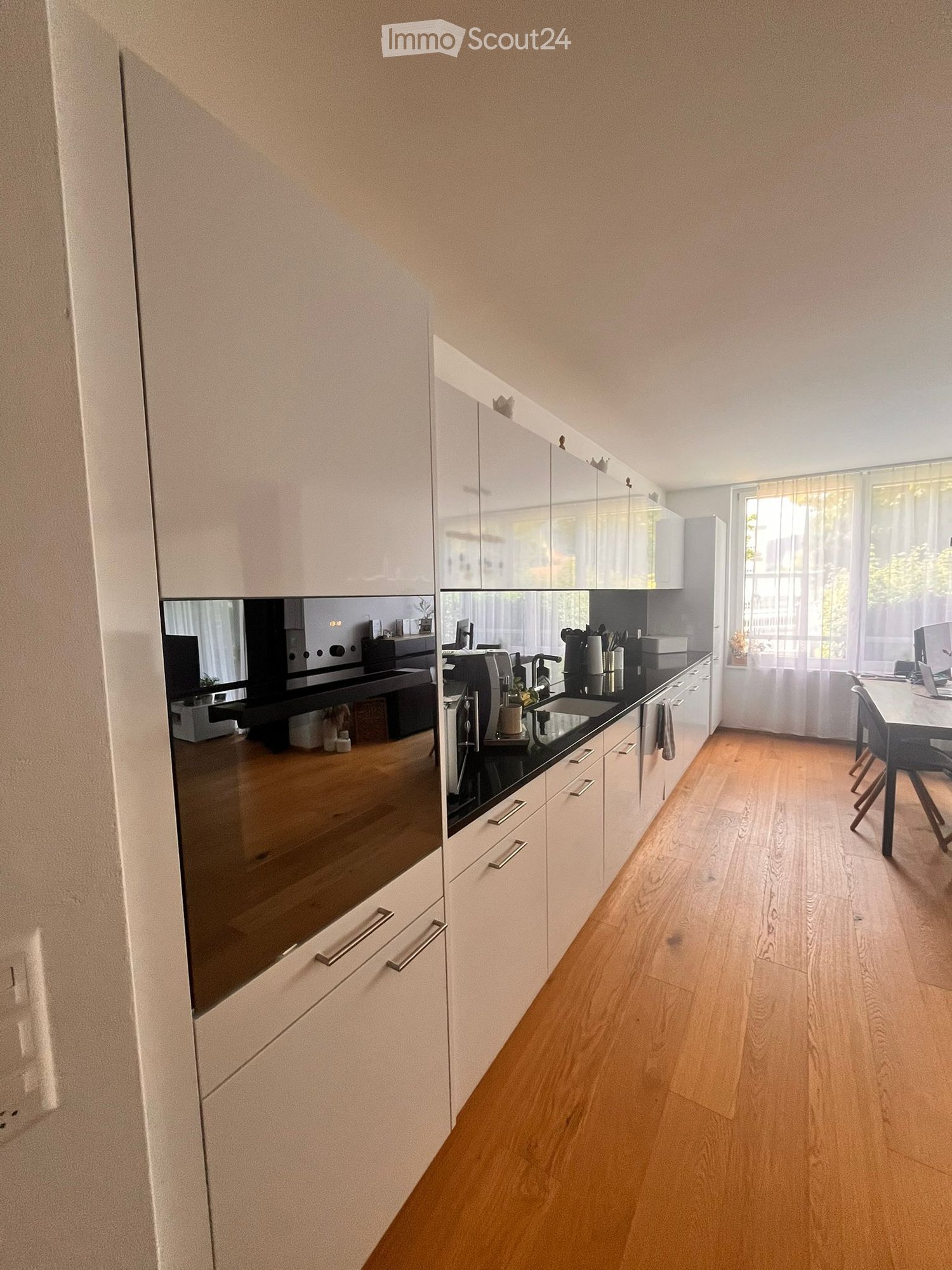 Modern kitchen with white cabinets, wooden flooring, and a black countertop. Includes a sink, coffee maker, and a small dining area with a table and chairs.