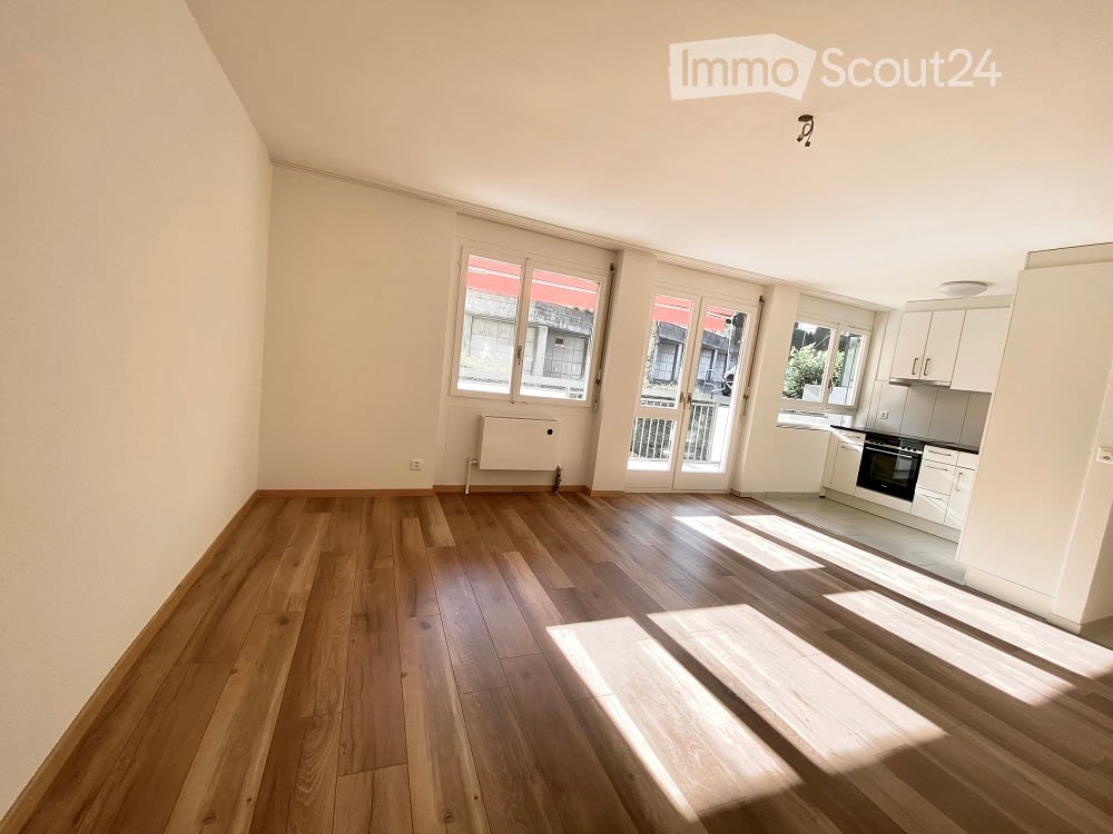 Bright, open-plan living and dining area with wooden flooring, large windows, and a small kitchen area featuring white cabinets and an oven. The space includes a balcony accessible through glass doors.