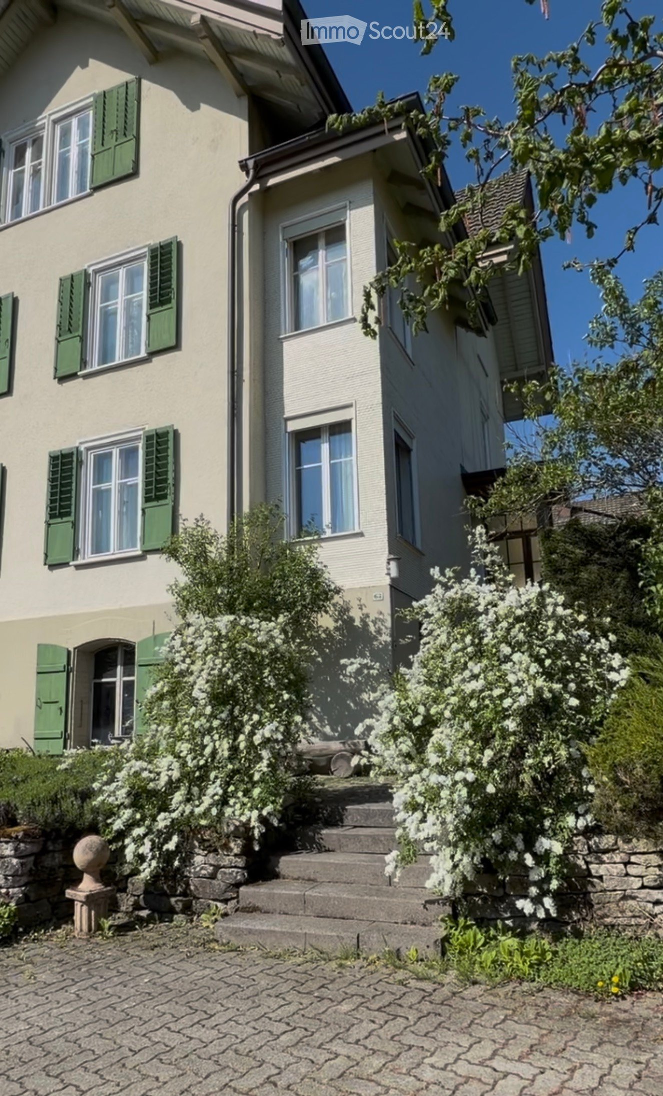 Two-story house with light-colored walls, green shutters, and multiple windows. The entrance features stone steps leading up to a door, surrounded by greenery and flowering bushes.