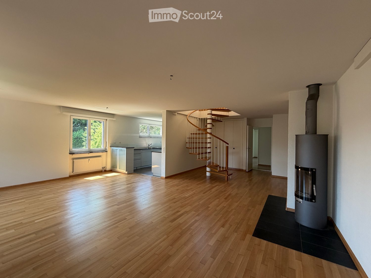 Bright, open-plan space with wooden flooring, a spiral staircase, a kitchen area, and a wood-burning stove. The room features large windows and neutral-colored walls.