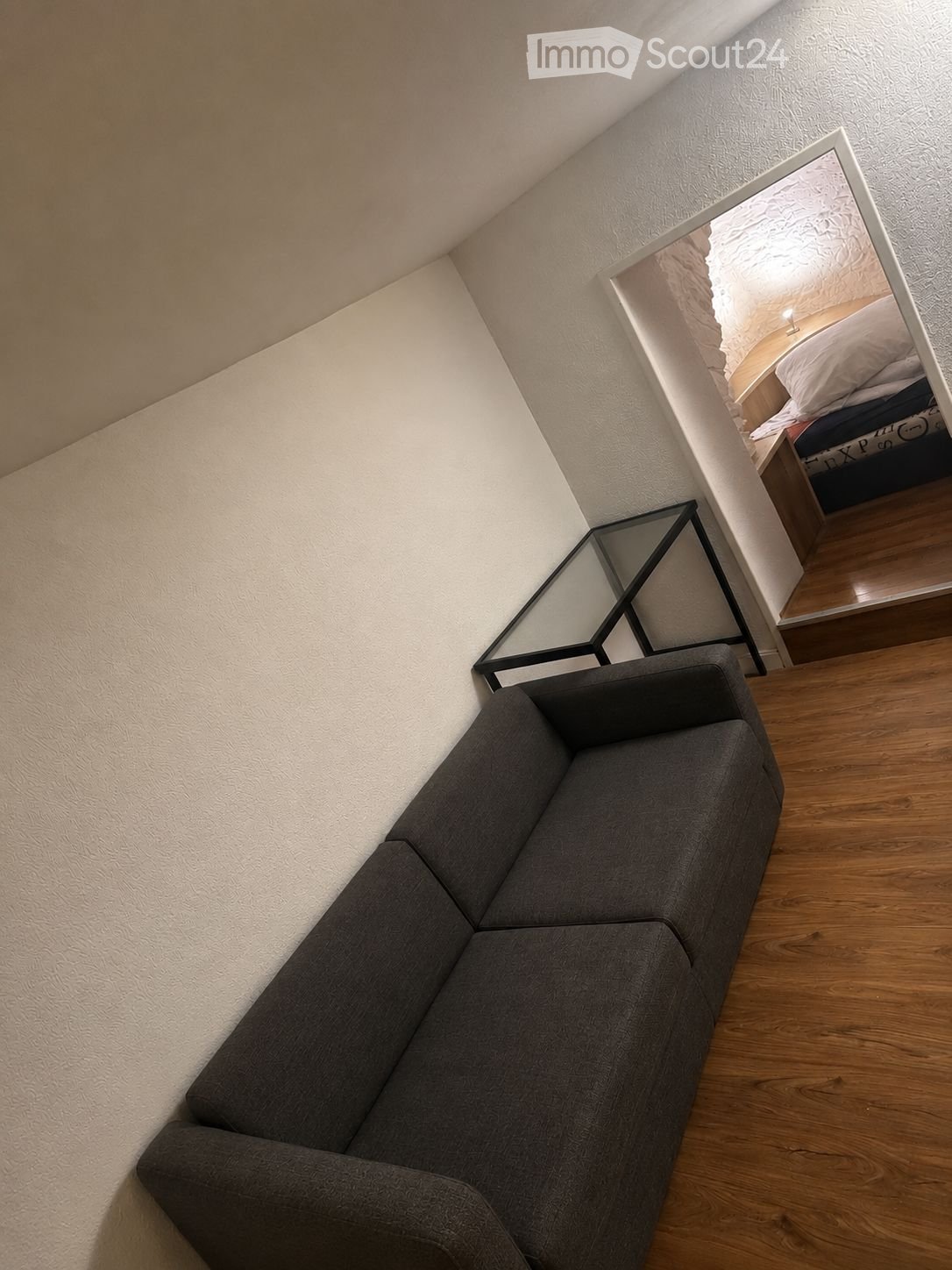 A modern living room with a grey sectional sofa, a sleek black glass table, and a large mirror reflecting a bedroom area with a bed and nightstand.
