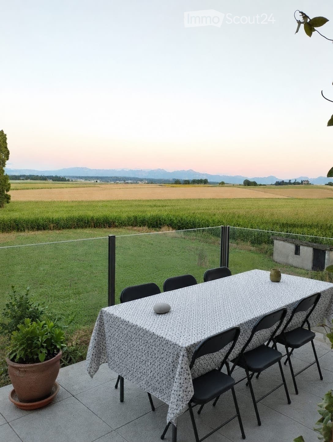 Balcony with a dining table and chairs, overlooking a green field and distant mountains.