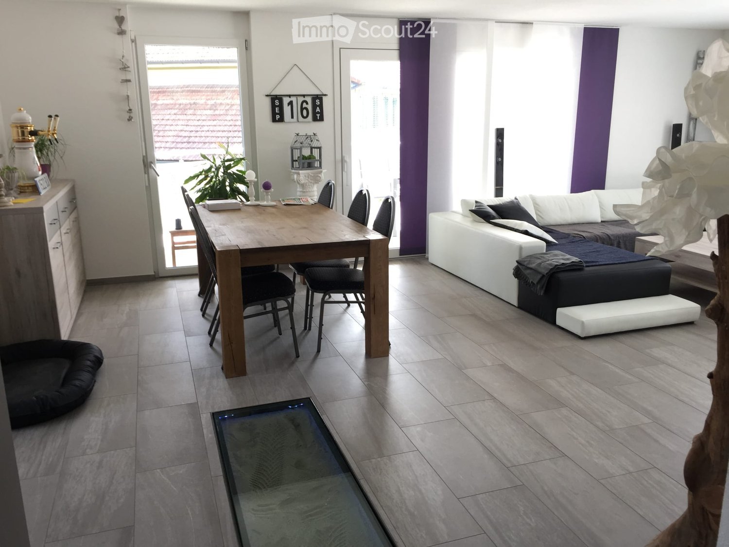 Open-plan living and dining area with a white sofa, wooden dining table, chairs, a small stool, and a plant near the window.