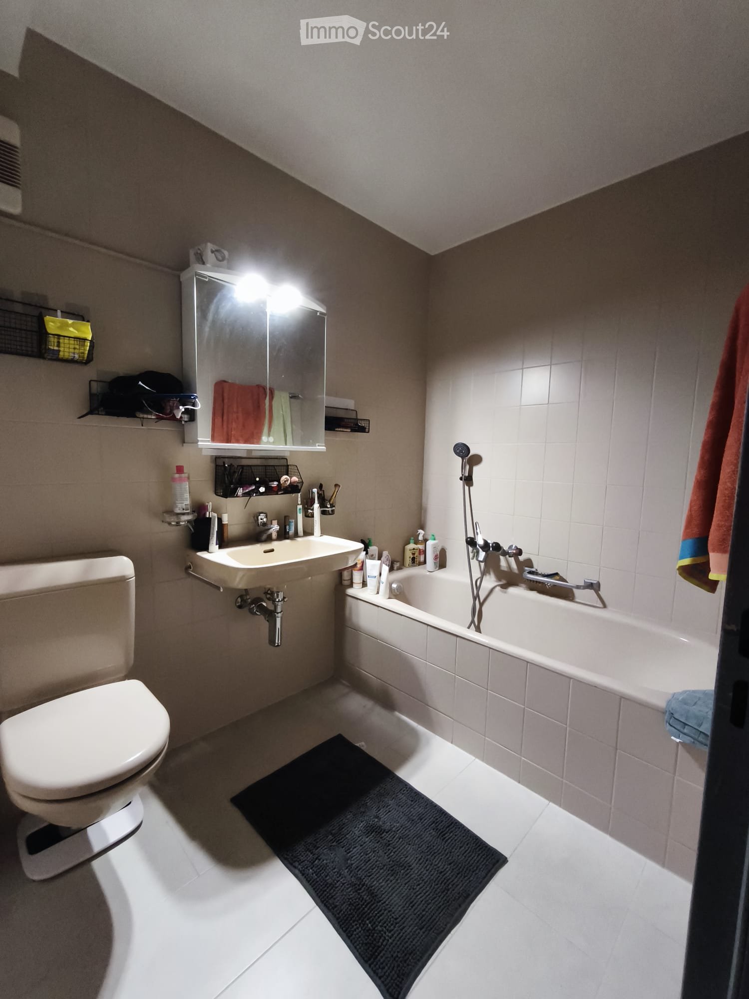 Bathroom with a toilet, sink, bathtub, mirror, and towel. The room features white tiles and a black bathmat on the floor.