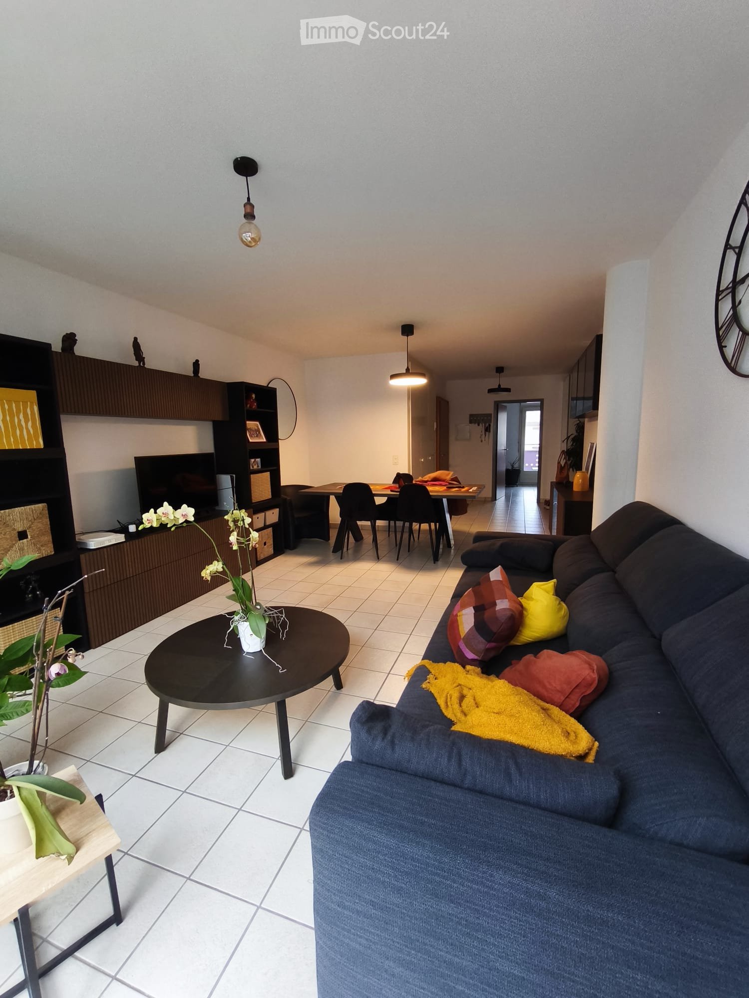Modern living room with a dark blue sectional sofa, coffee table, dining table with chairs, television, bookshelf, and decorative plants.