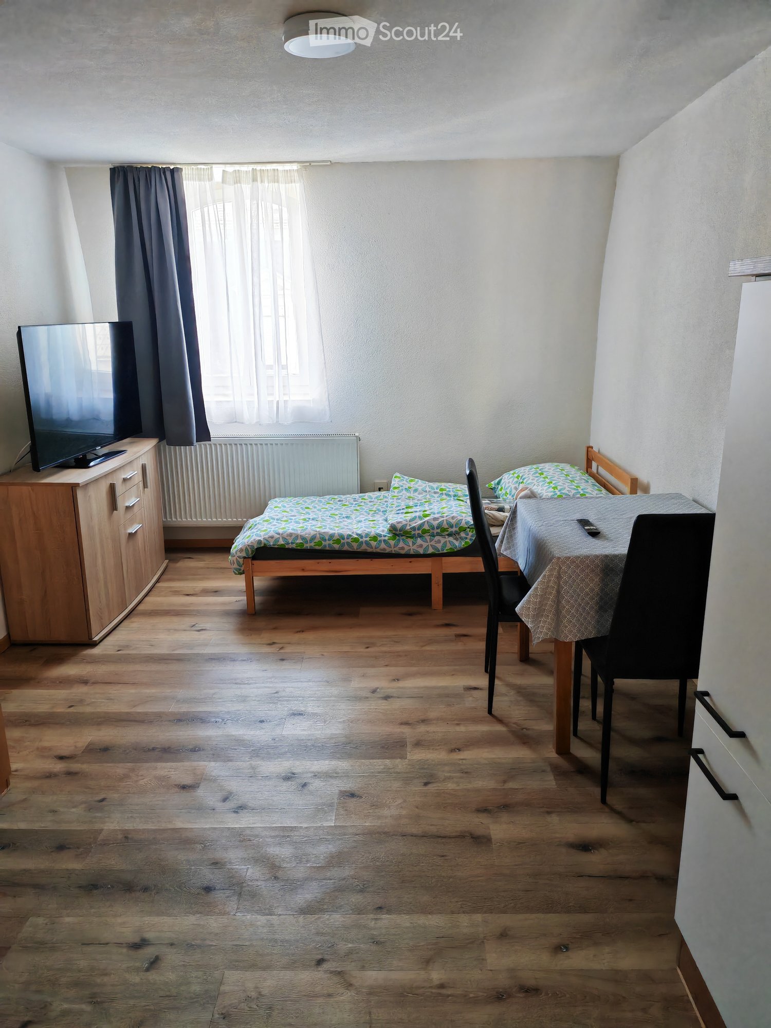 A small, furnished room with a wooden floor, a single bed with patterned bedding, a small table with chairs, a television on a wooden cabinet, and a window with curtains.