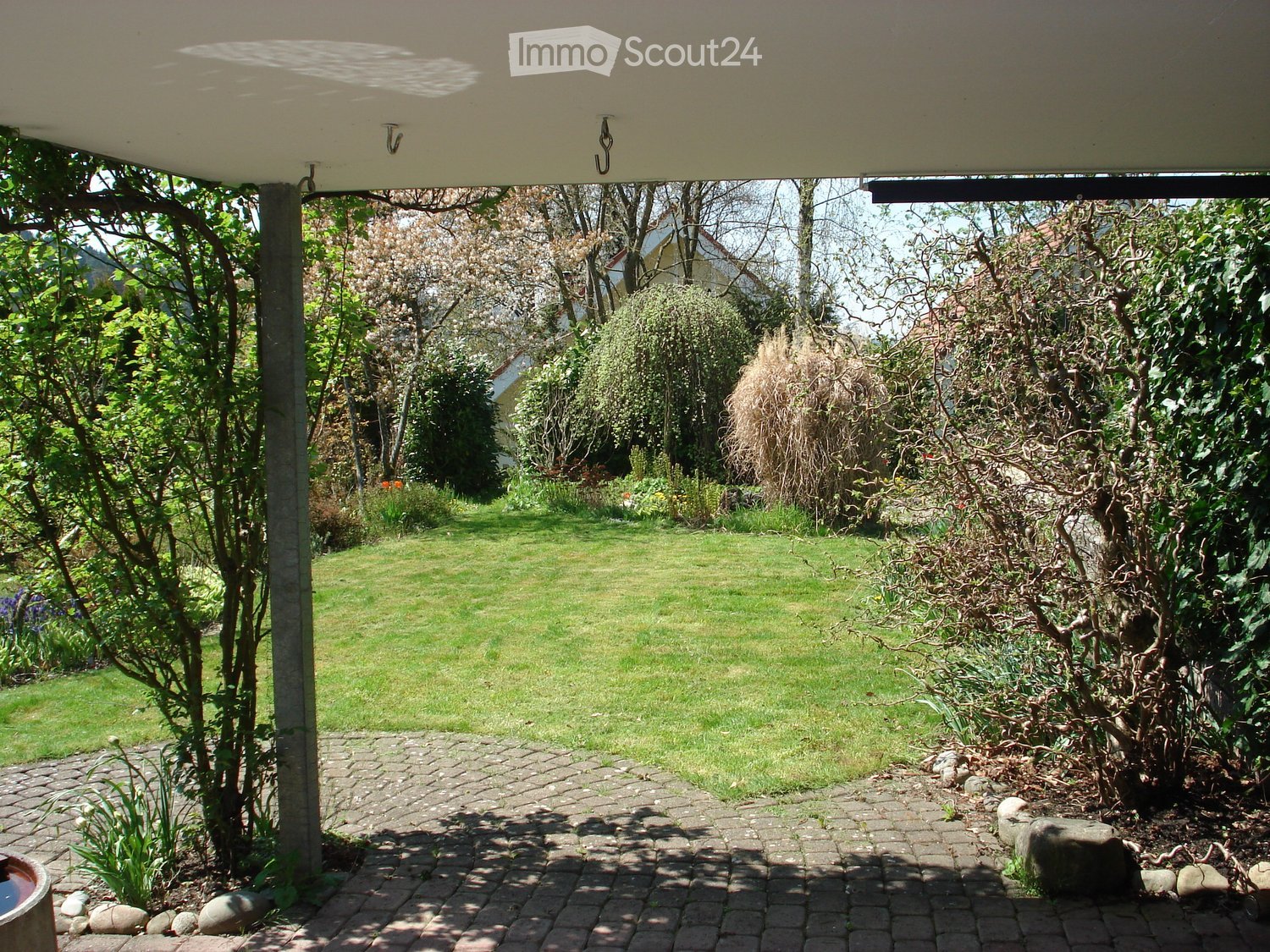 Well-maintained garden with a paved pathway, grassy lawn, trees, and shrubs. A covered patio area is visible in the background.
