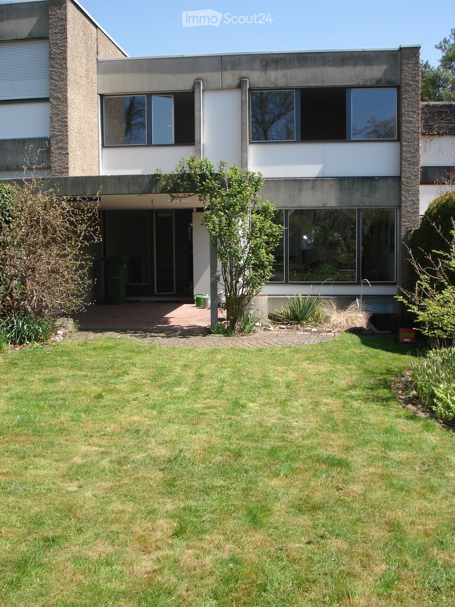 Modern two-story house with large windows, concrete facade, and a well-maintained lawn in front.