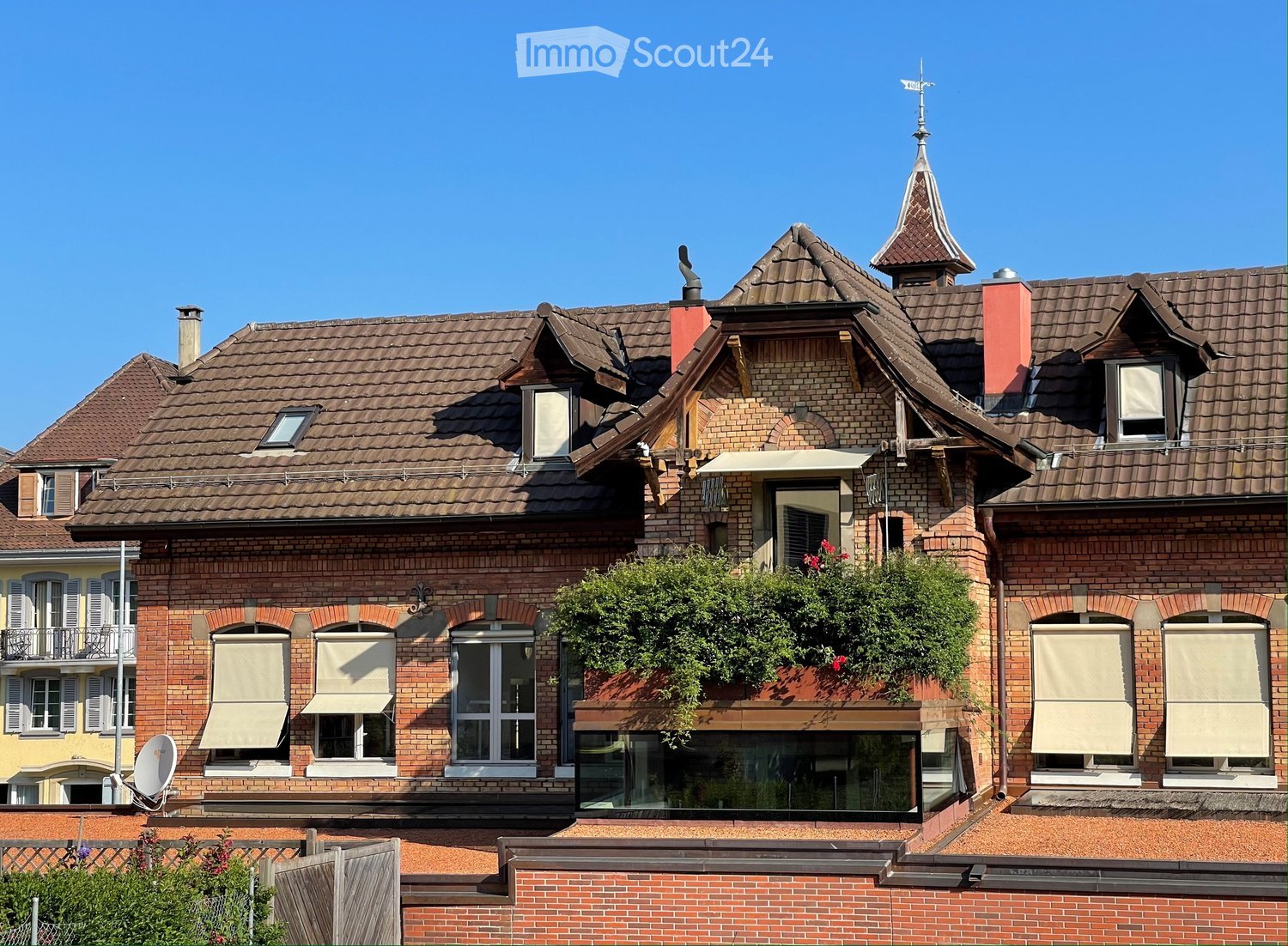 A multi-story brick house with a tiled roof, multiple chimneys, and several windows with shutters. The building features a balcony with greenery and a satellite dish on the roof.