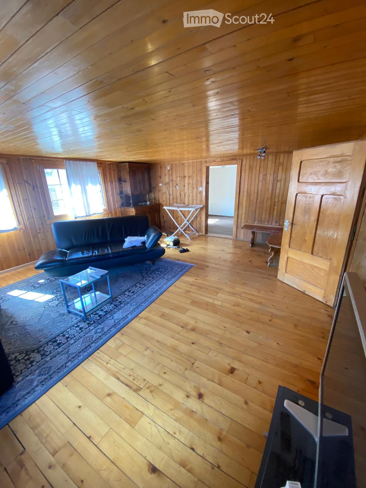 A spacious living room with wooden floors and walls, featuring a black leather sofa, a glass coffee table, a bench, and a small table with chairs.
