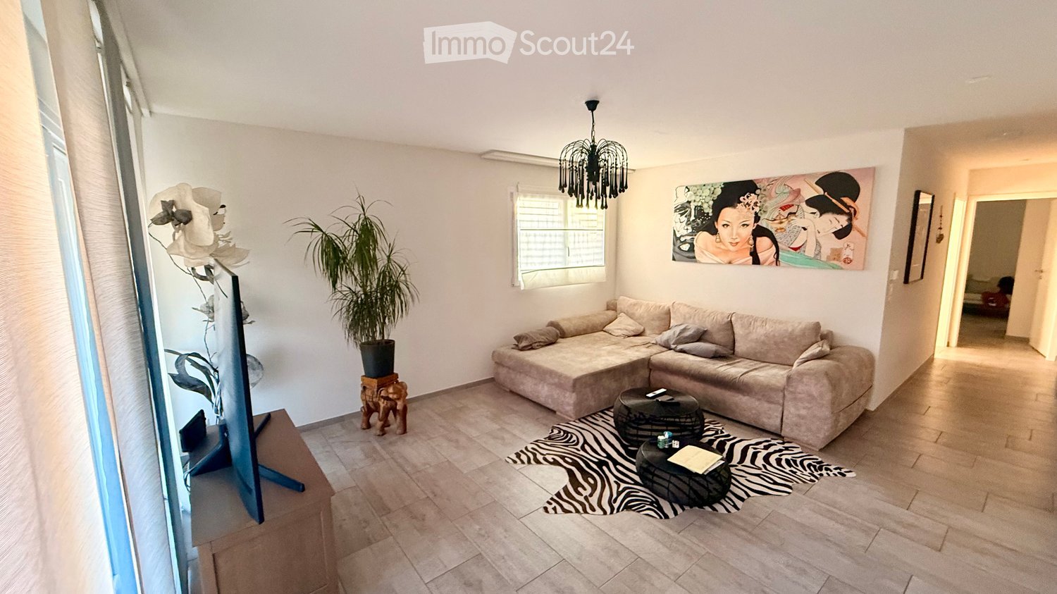 Modern living room with a beige sectional sofa, coffee table, zebra print rug, and decorative plant. Features a large window, a chandelier, and a colorful artwork on the wall.