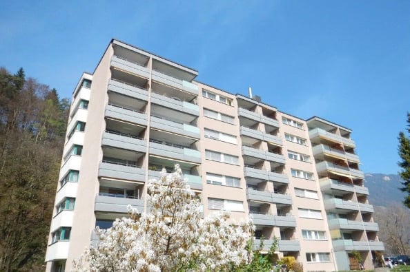 Multi-story residential building with multiple balconies and windows, surrounded by greenery and trees.