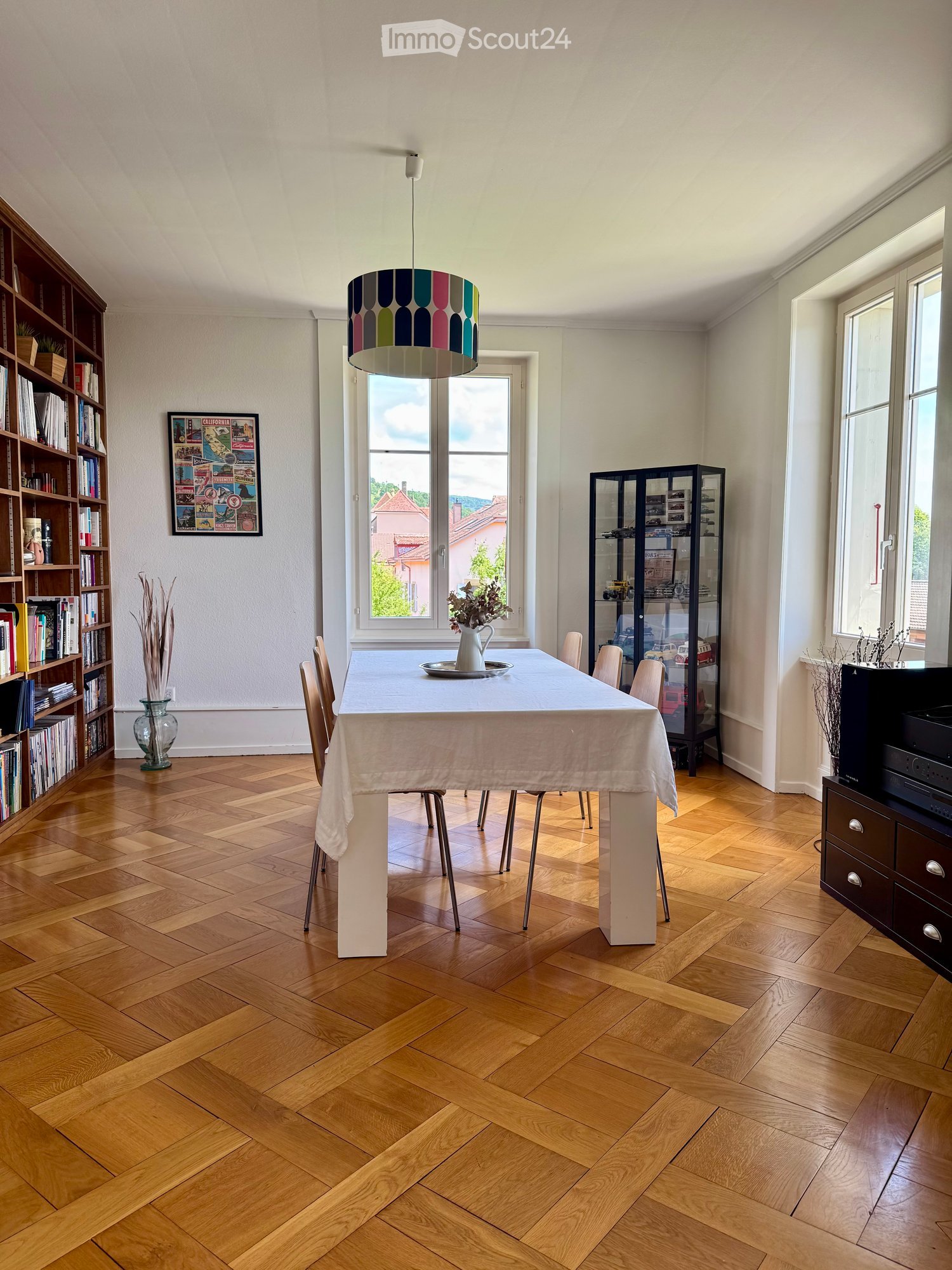 Bright kitchen with wooden herringbone flooring, large windows, dining table with chairs, bookshelf, decorative vase, and a colorful pendant lamp.