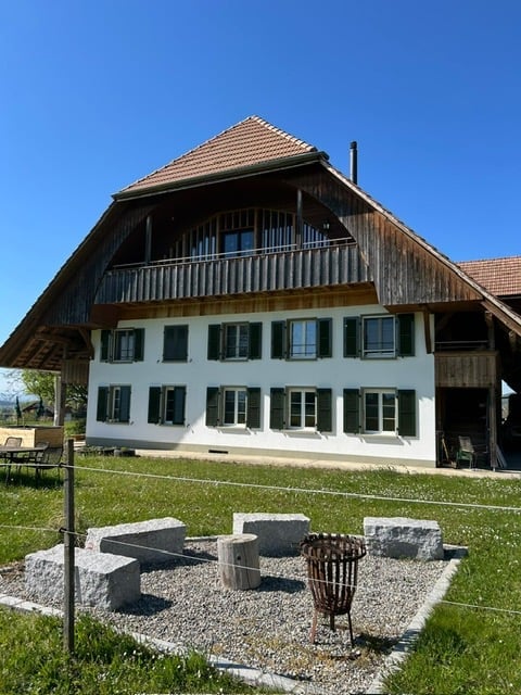 House. Traditional two-story house with white walls, dark green shutters, and a wooden balcony on the upper floor. The roof is red-tiled with a prominent wooden overhang. The surrounding area includes a lawn, a small seating area with stone blocks and a m
