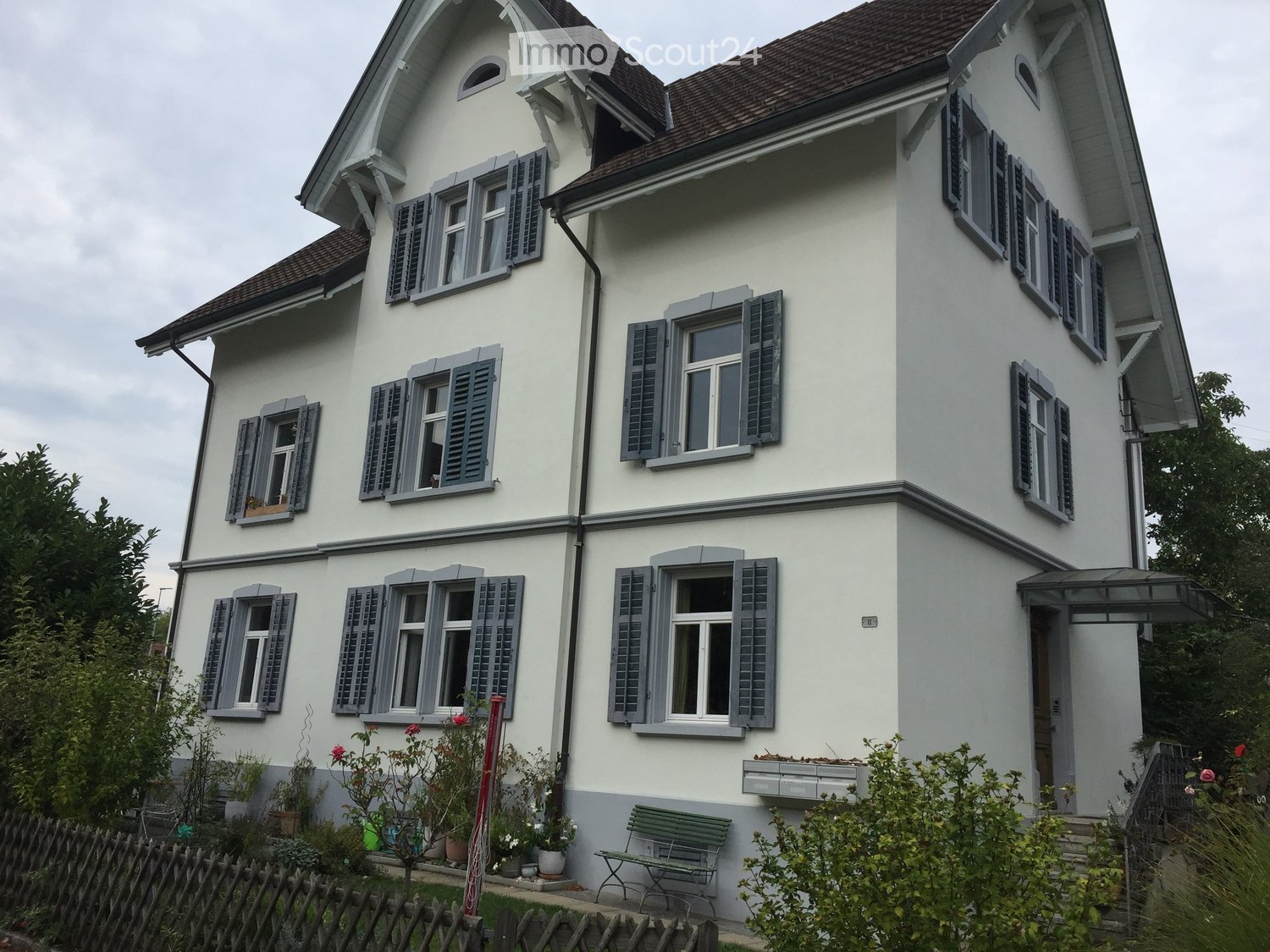 House. Two-story house with white exterior, grey shutters, multiple windows, and a small garden area with a bench and potted plants.