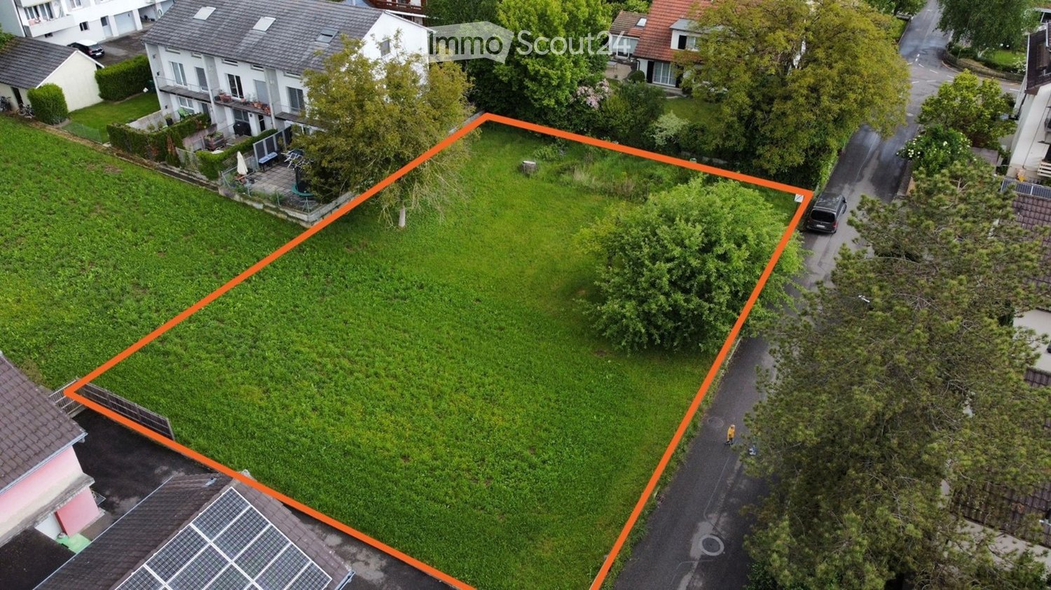 Aerial view of a large, well-maintained lawn surrounded by neighboring houses and trees. The lawn is enclosed by a fence and features a small pathway leading to the street.