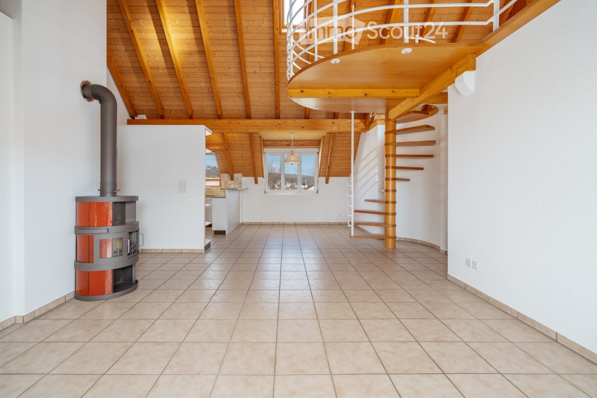 Bright hallway with wooden ceiling beams, tiled floor, and a modern wood-burning stove on the left side. A spiral staircase leads to an upper level.