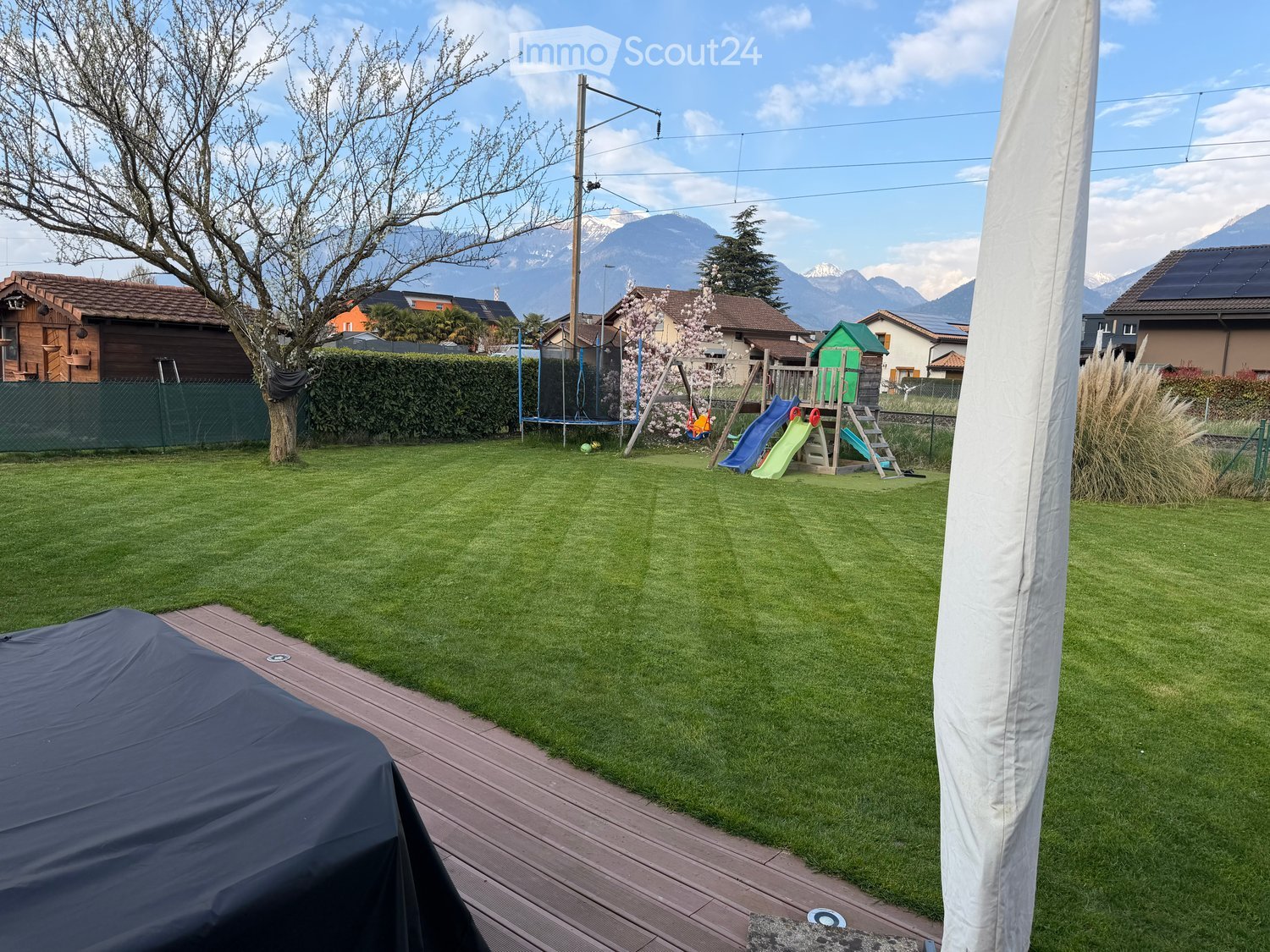 Well-maintained lawn with a wooden deck, covered grill, and a children's play area including a slide and swing set. The background features a hedge, trees, and distant mountains.