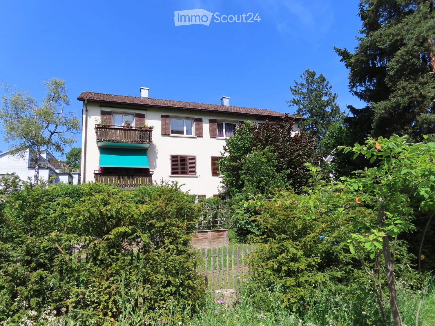 Two-story house with white walls, brown shutters, and a green balcony awning. The house is surrounded by lush greenery and trees.