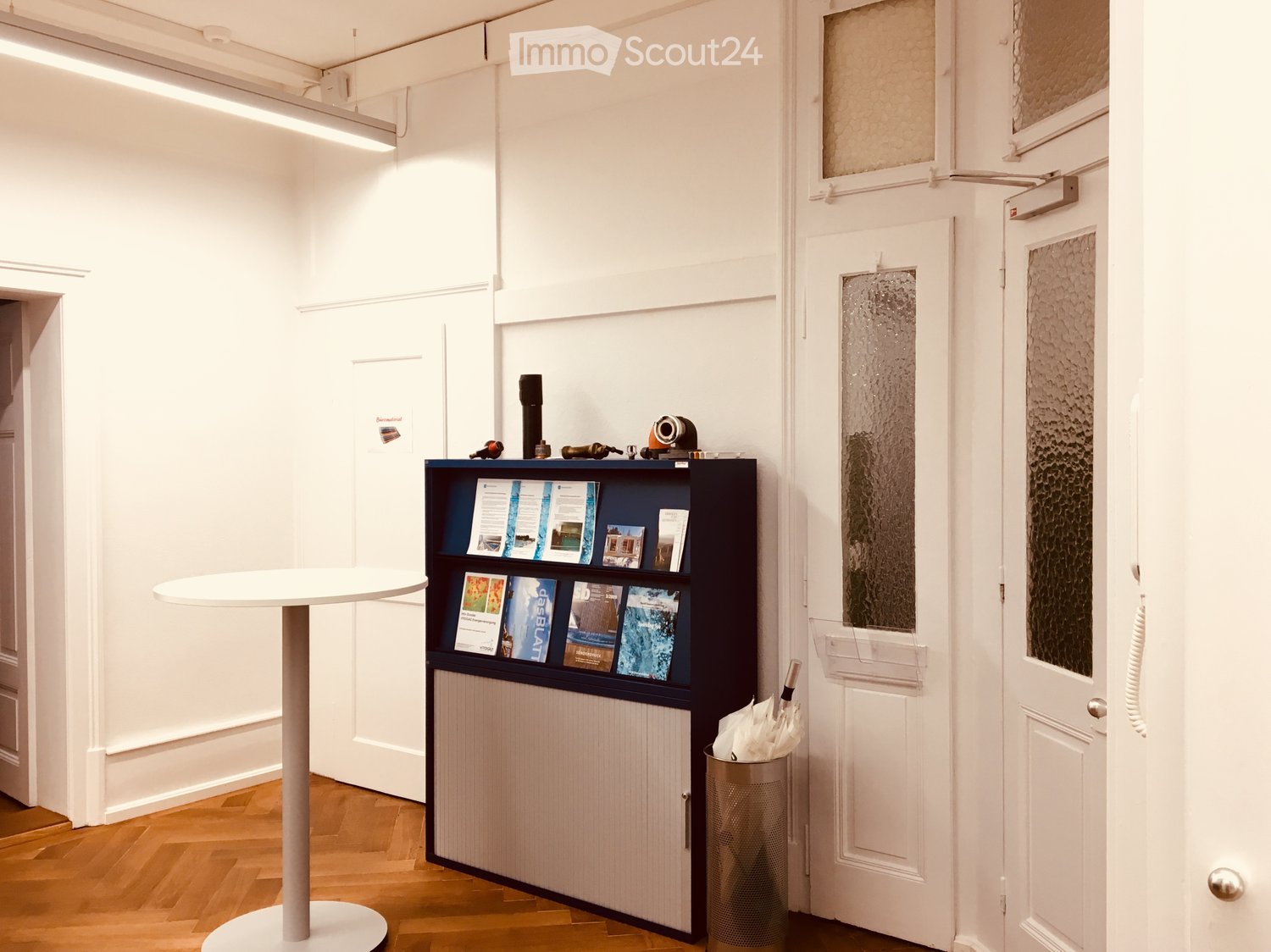 A modern, minimalistic hallway or reception area with a white table, a dark wooden shelf holding brochures, and a small umbrella stand. The floor is wooden, and the walls are white with framed artwork.