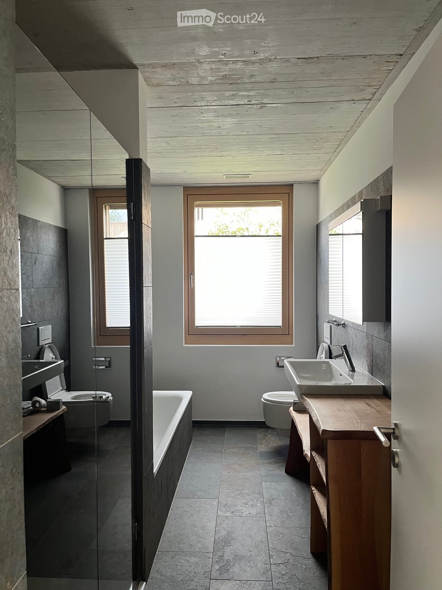 Modern bathroom with a wooden vanity, white sink, bathtub, toilet, and large windows providing natural light.