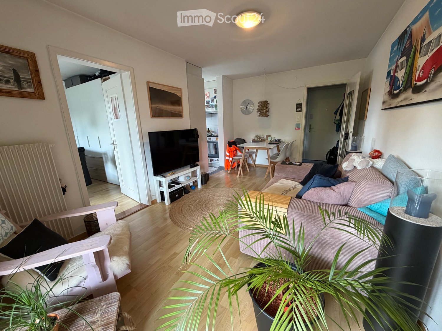 A cozy living room with a sofa, armchair, coffee table, television, and a small dining area visible in the background. The room features wooden flooring, a round rug, and several decorative items including a plant and artwork on the walls.