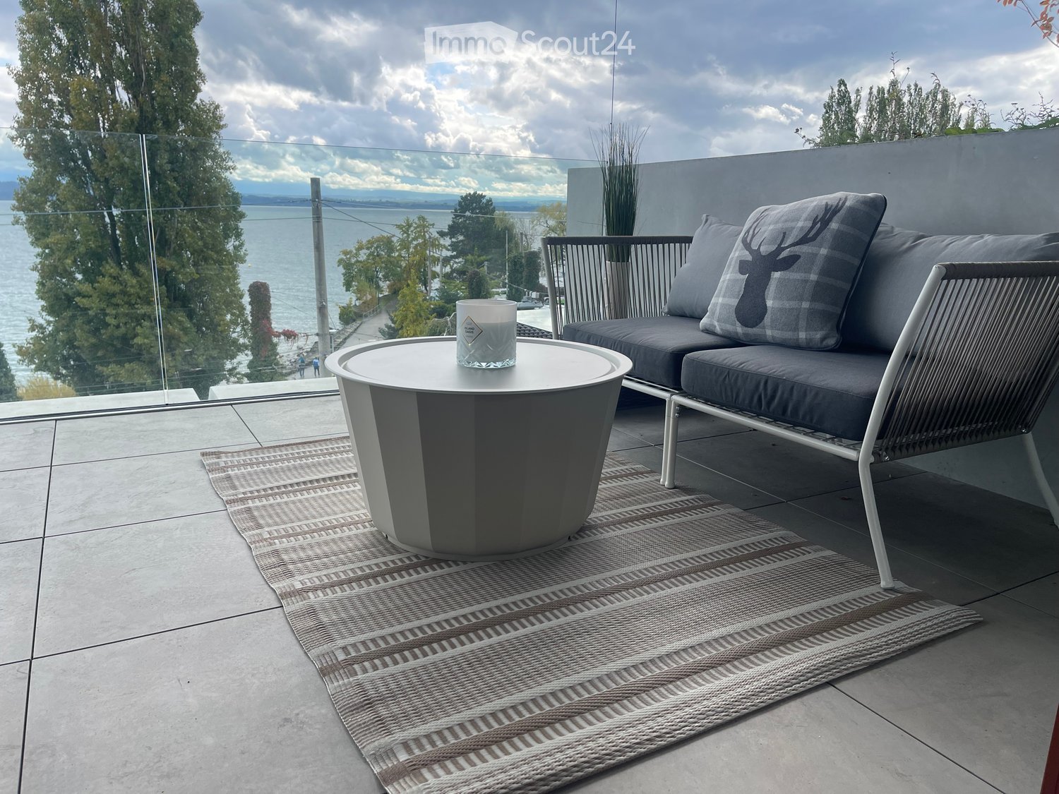 Outdoor balcony with sofa, round table, rug, glass railing, lake view, and lush greenery