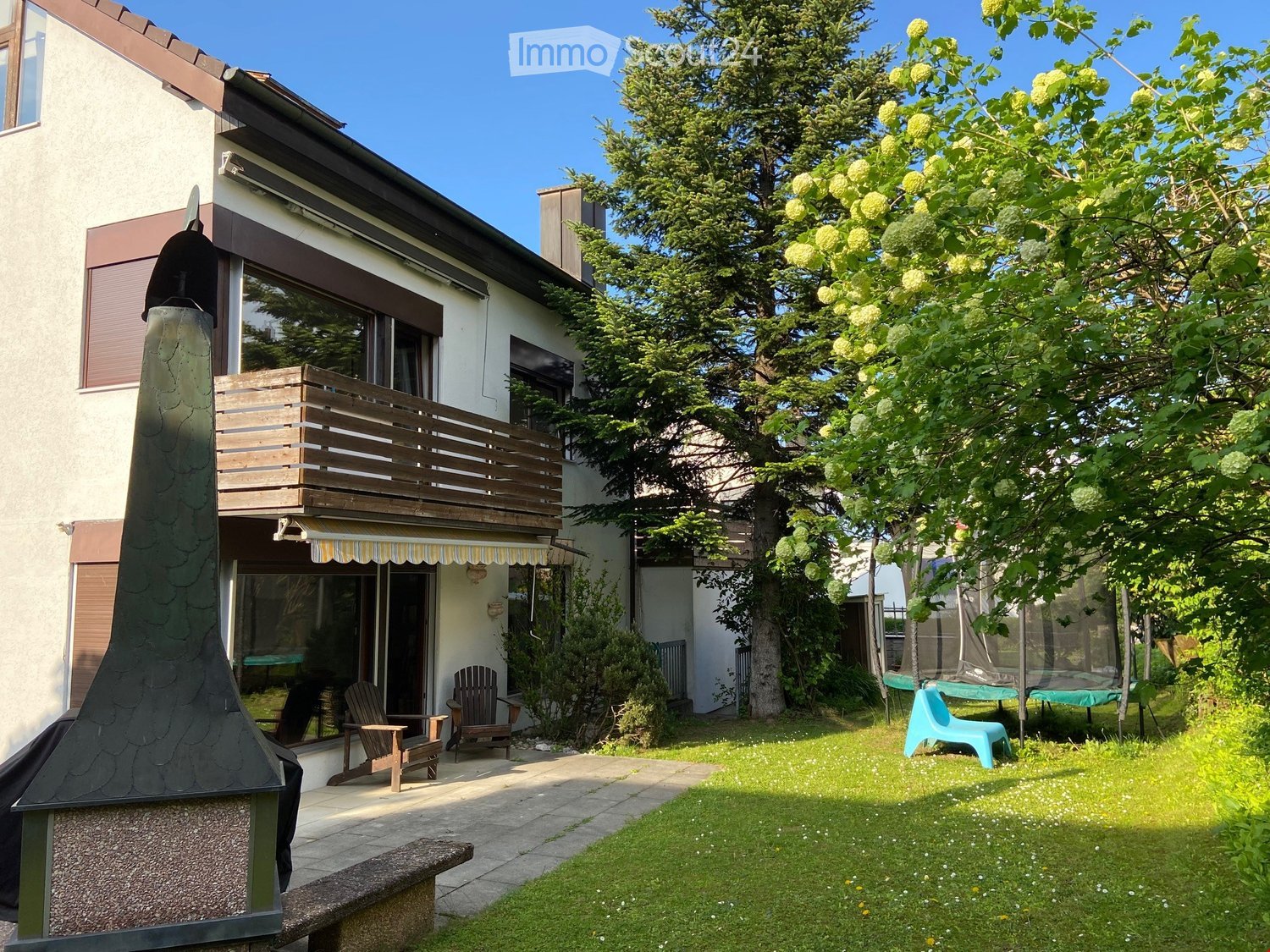 Two-story house with a balcony, wooden deck, and outdoor seating area. The garden includes a lawn, trees, and patio furniture.