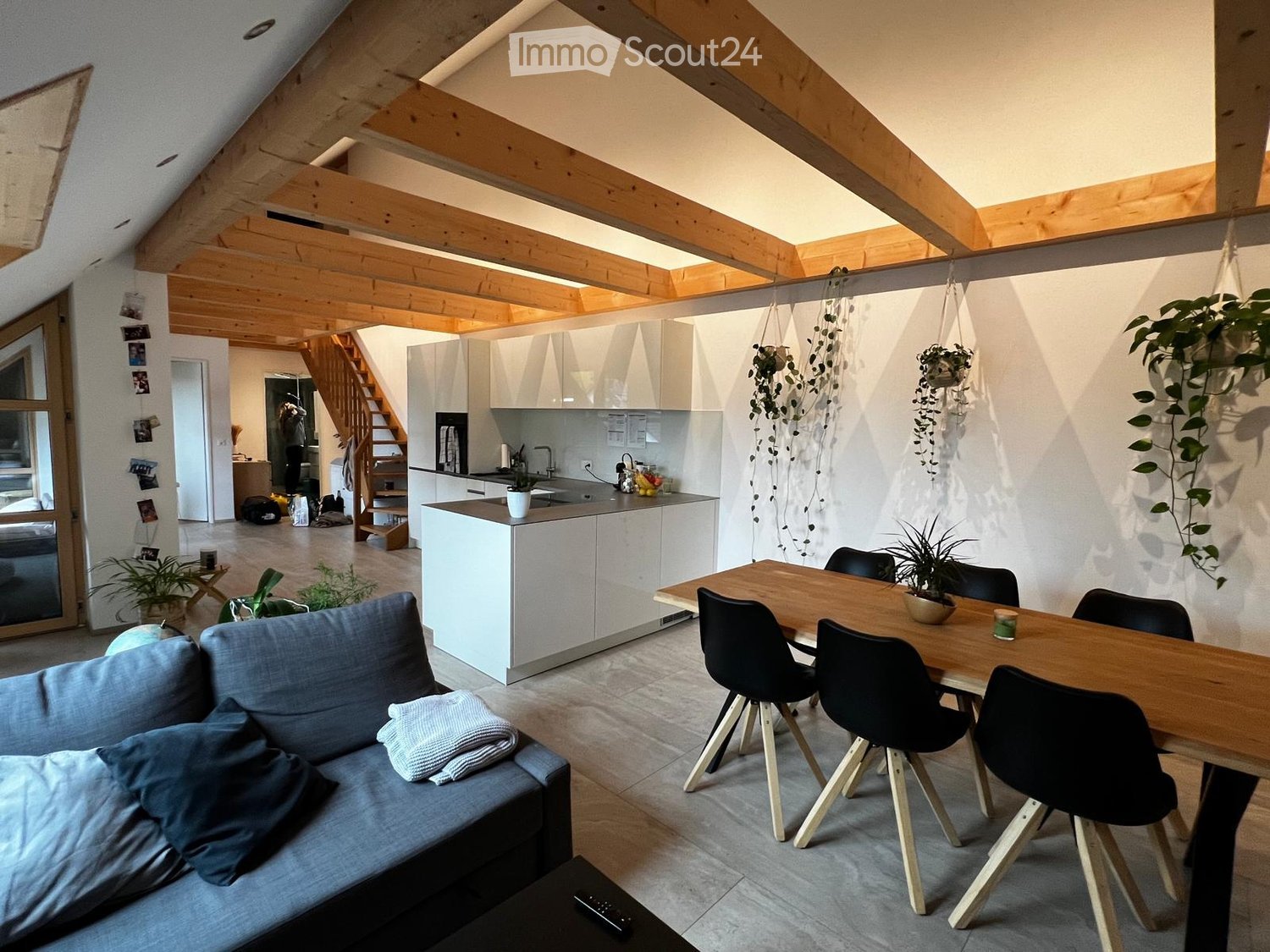 Open-plan living and dining area with exposed wooden beams, grey sofa, wooden dining table with black chairs, modern white kitchen, hanging plants, and a staircase leading to another level.