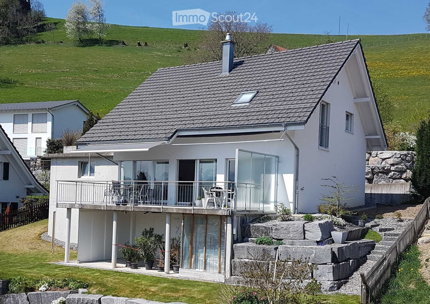 A modern two-story house with a grey tiled roof, white exterior, and a spacious balcony with glass railings. The house is surrounded by a well-maintained garden with potted plants and a stone wall.