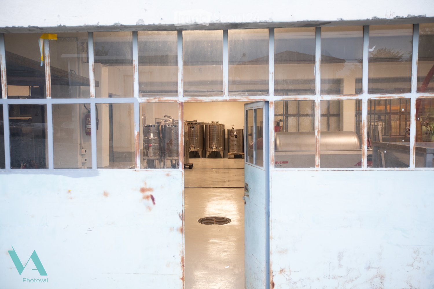 View through a large industrial-style window into a spacious, well-lit interior with stainless steel tanks and equipment, likely a winery or brewery.