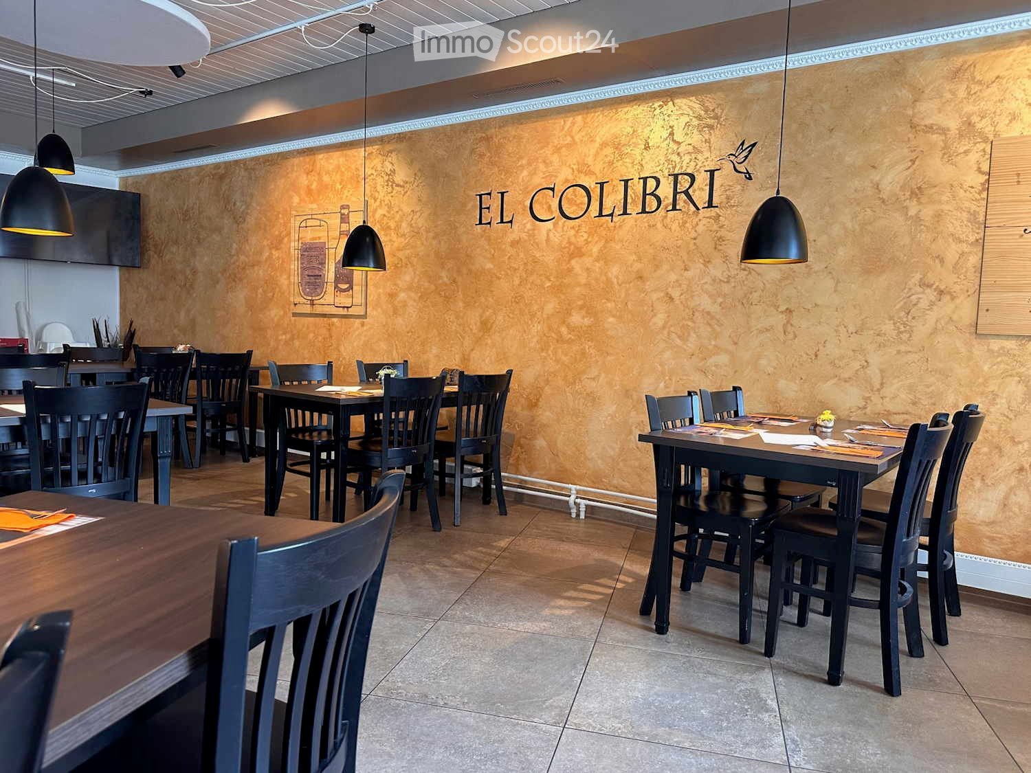 Restaurant interior with dark wooden tables, black chairs, pendant lights, and a textured wall with the name "EL COLIBRI".