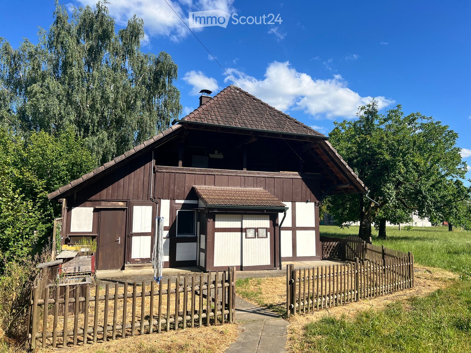 Traditional wooden house with a pitched roof, white and brown exterior, surrounded by a wooden fence and trees.