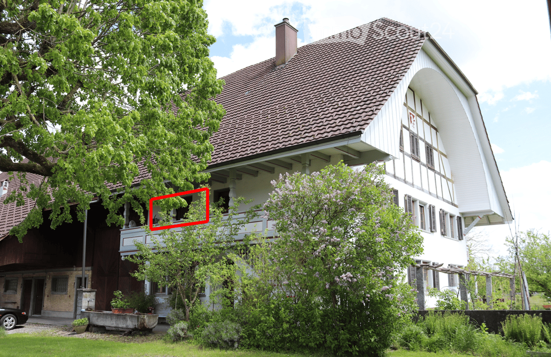 Two-story house with a tiled roof, white exterior, and a balcony on the upper level. The house is surrounded by greenery, including trees and flowering shrubs.