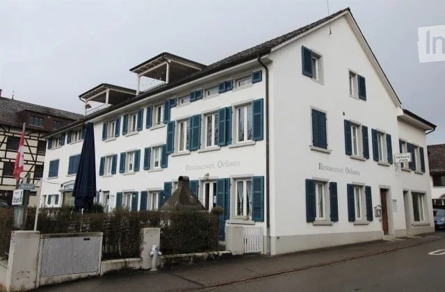 A two-story white building with blue shutters and a restaurant sign reading "Restaurant Ochsen." The building has multiple windows and a small garden area in front.