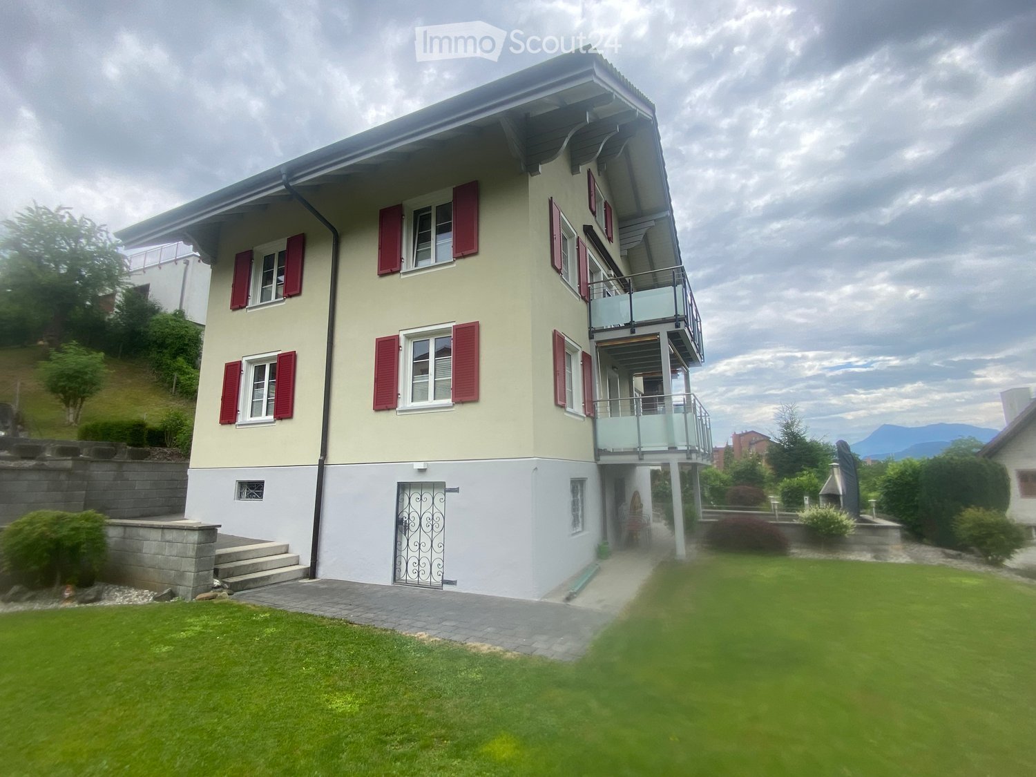 Two-story house with beige walls, red shutters, and a balcony on the upper level. The house features a well-maintained lawn, a paved pathway, and a stone retaining wall.