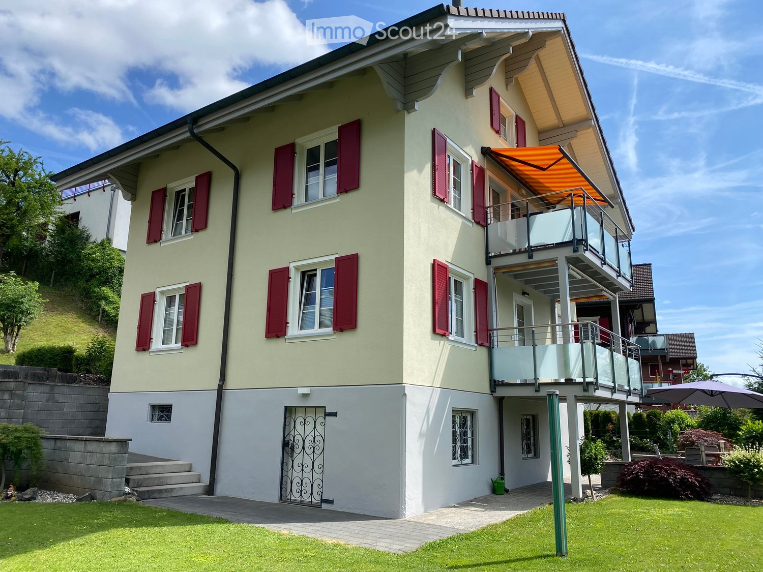 Two-story house with light yellow walls, red shutters, and a balcony with an awning. The house features a raised ground floor, a well-maintained lawn, and a small garden area with trees and shrubs.