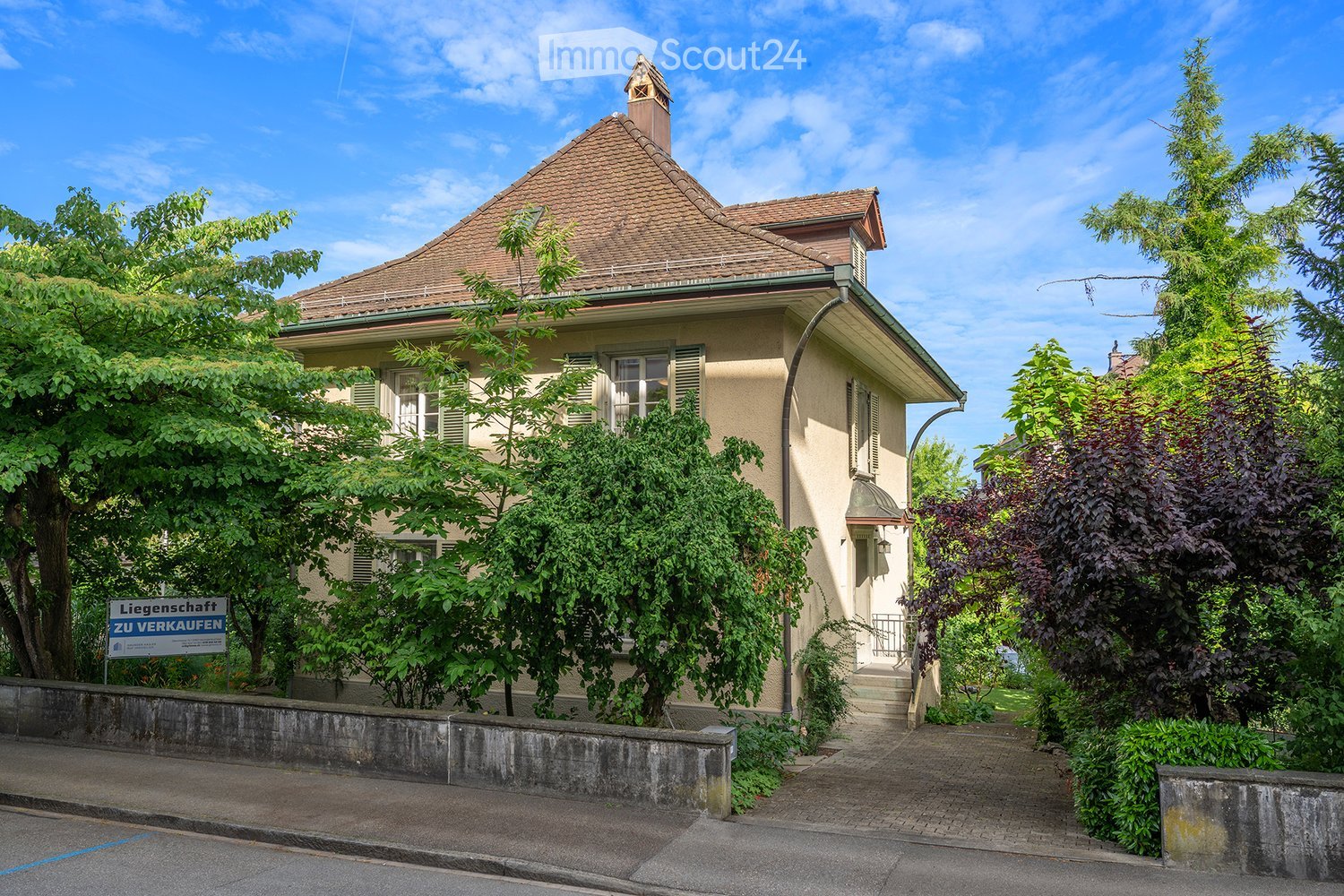 Two-story house with a tiled roof, multiple windows, and a small balcony. The house is surrounded by greenery and has a "Liegenschaft zu Verkaufen" sign indicating it is for sale.