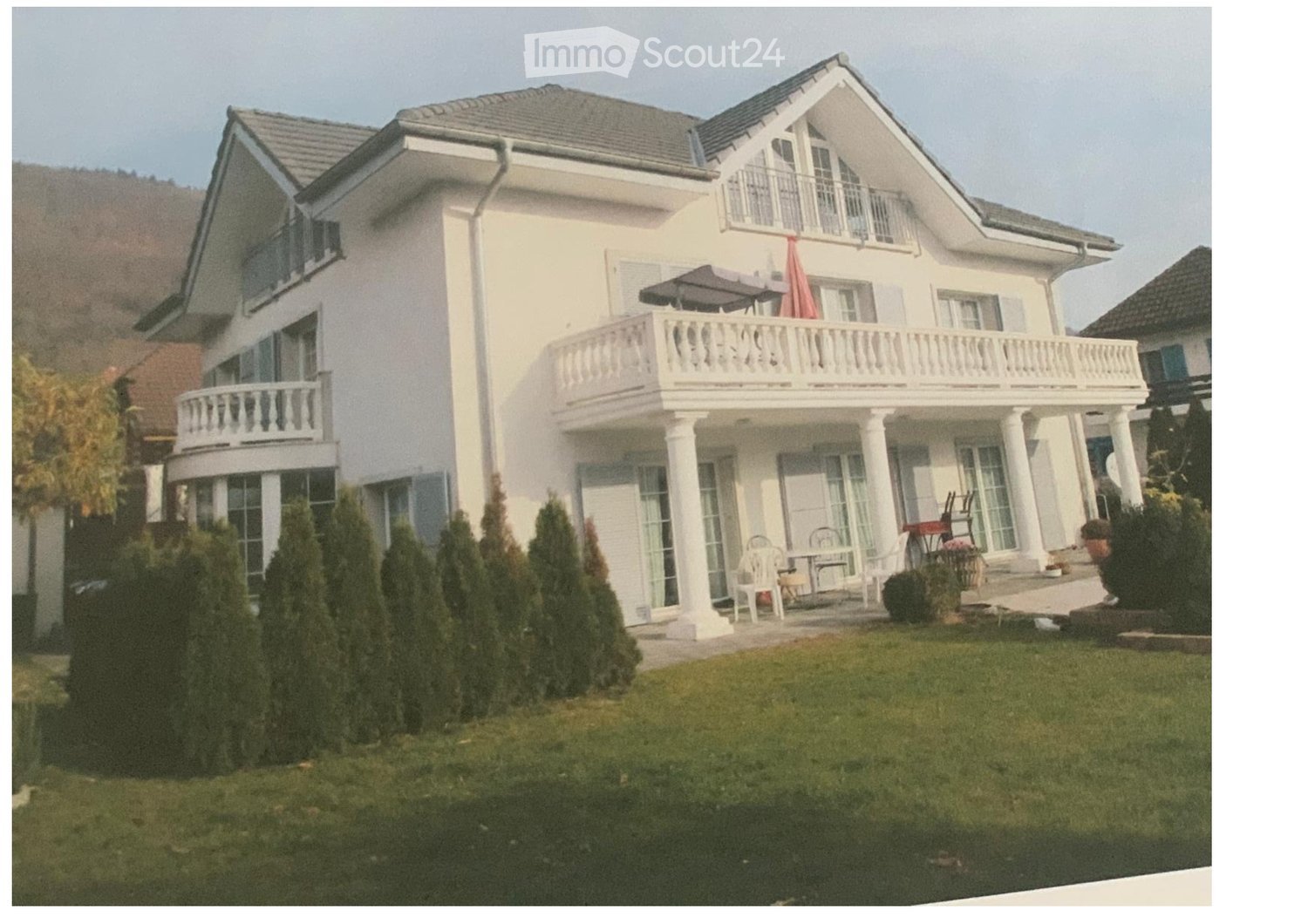 Two-story white house with multiple balconies, large windows, and a well-maintained lawn with trimmed bushes.