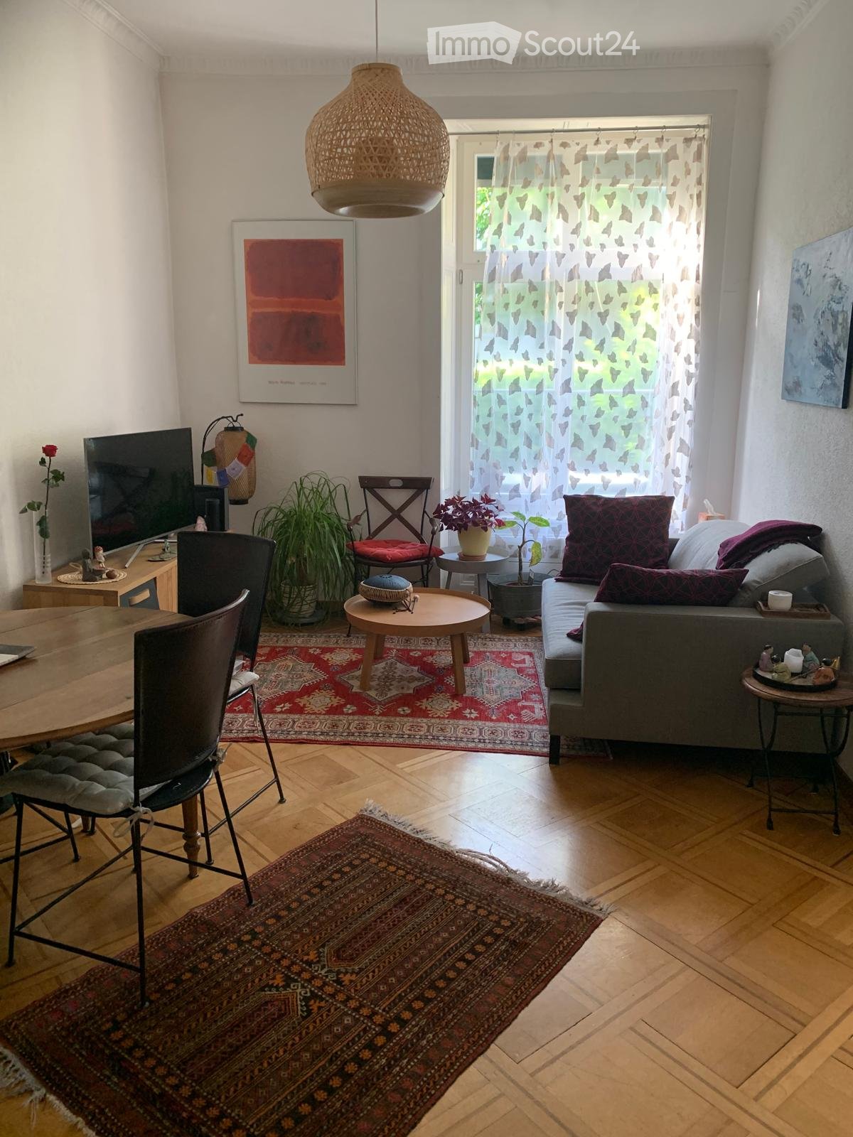Living room with wooden flooring, a sofa with pillows, a coffee table, a dining table with chairs, a television, and decorative items including potted plants and artwork.