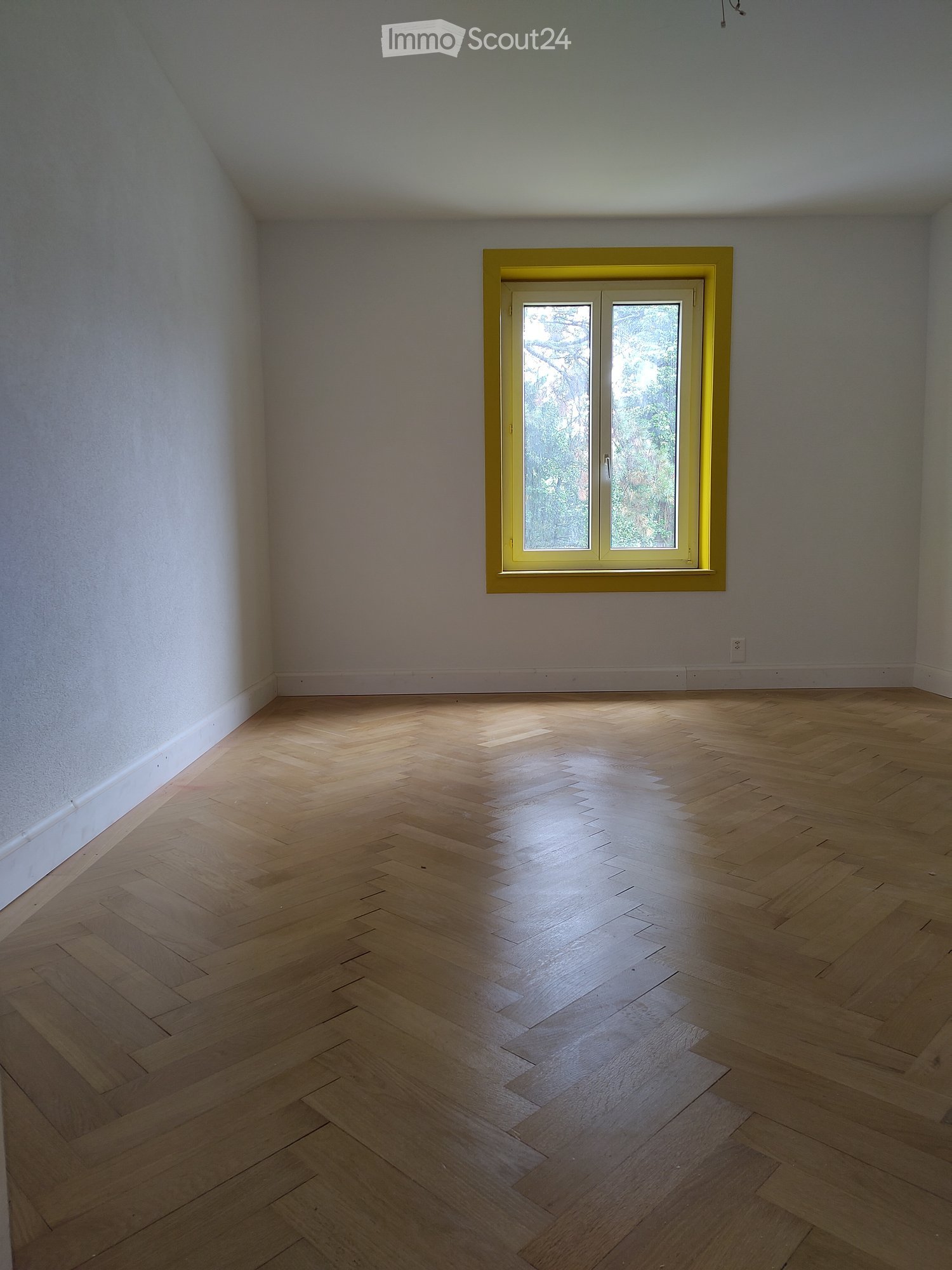 Empty room with wooden herringbone flooring and a single window with yellow frame, allowing natural light to enter.